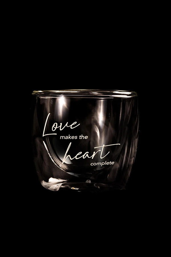 A clear glass espresso cup with black text reading 'Love makes the heart complete' printed on it.
